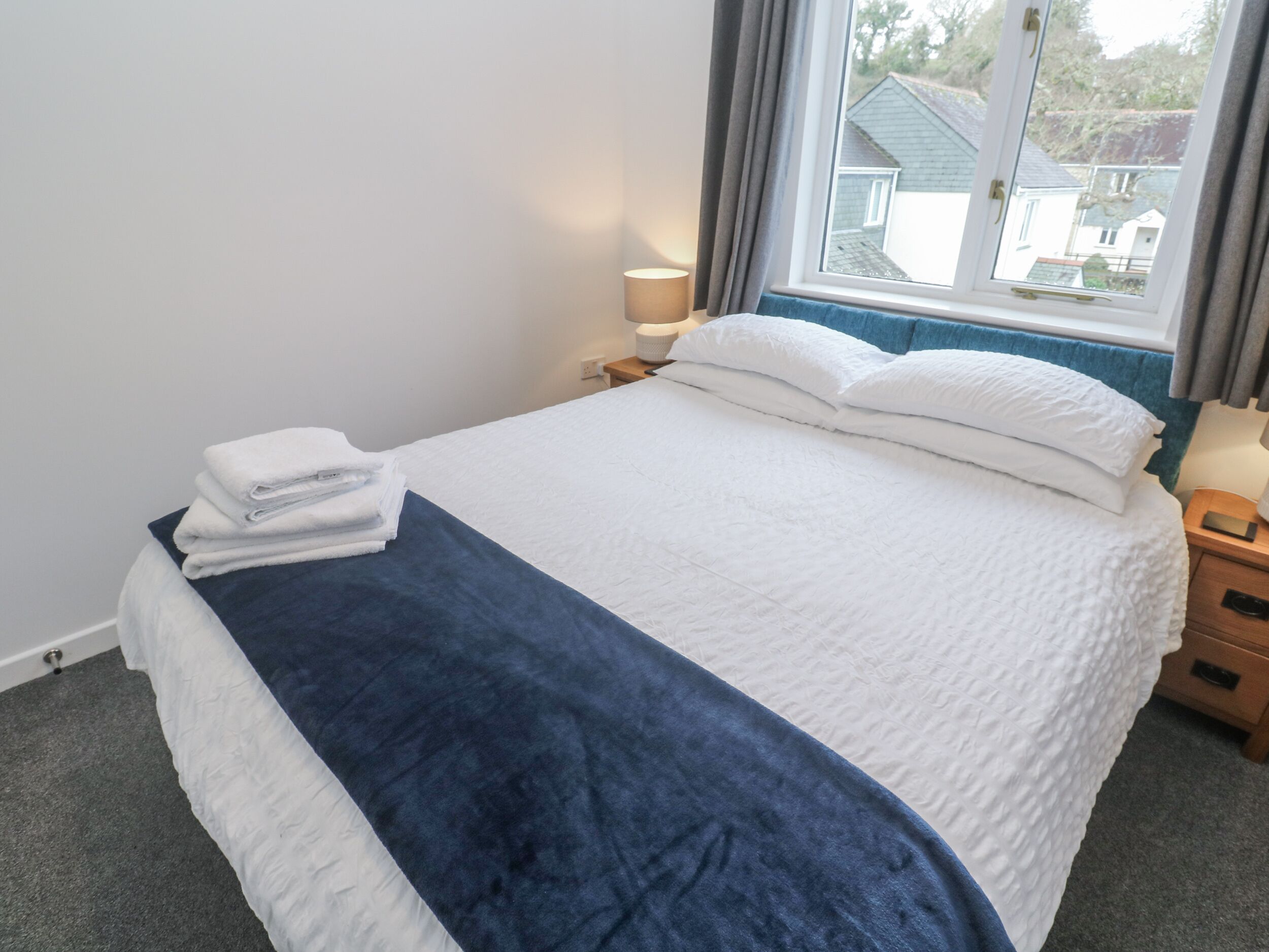 Una cama bien tendida con ropa de cama blanca y azul, una lámpara de noche y vistas a casas a través de una ventana.
