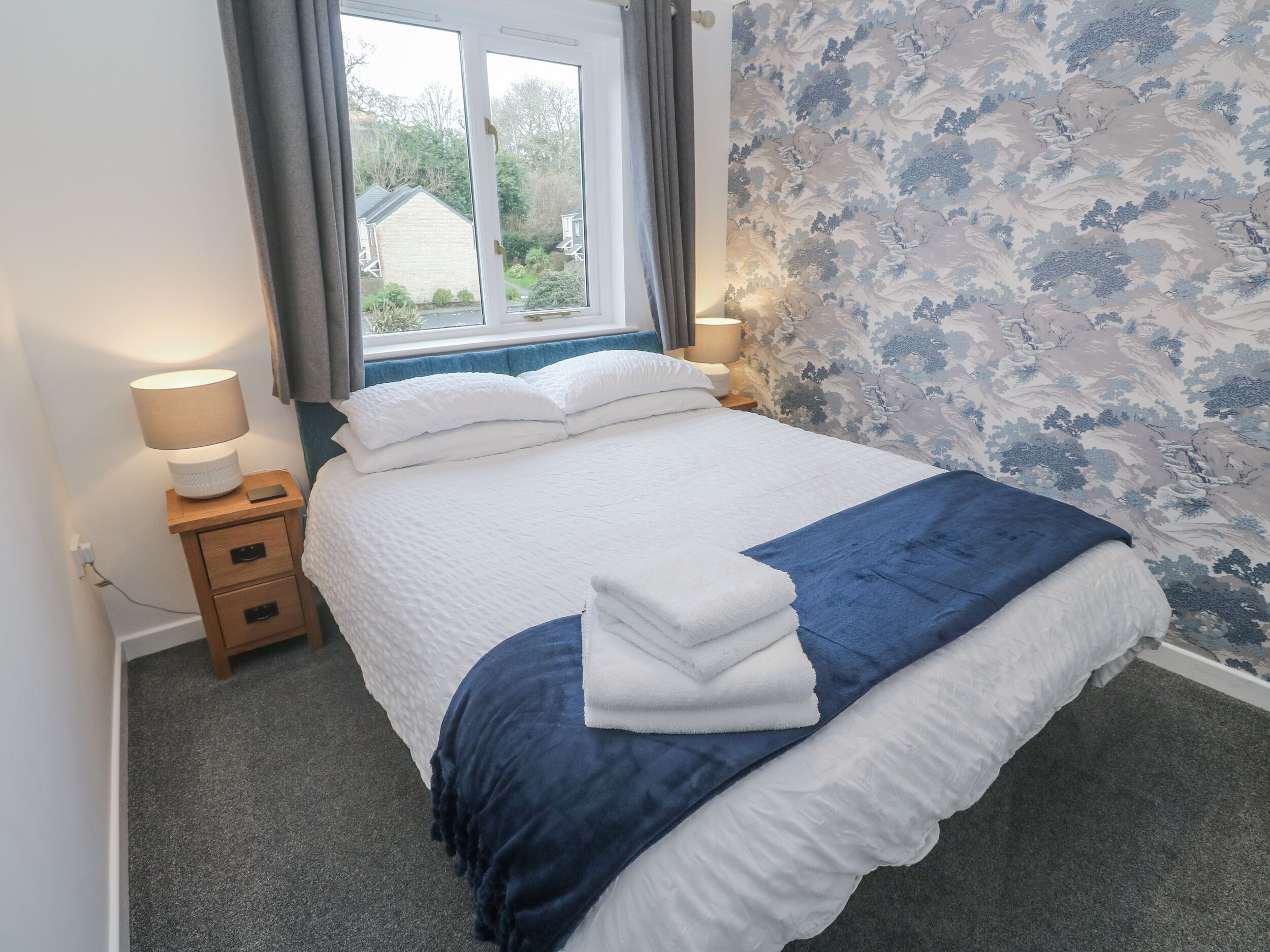 A neatly made bed with white and blue bedding, a bedside table with a lamp, and a window with curtains.