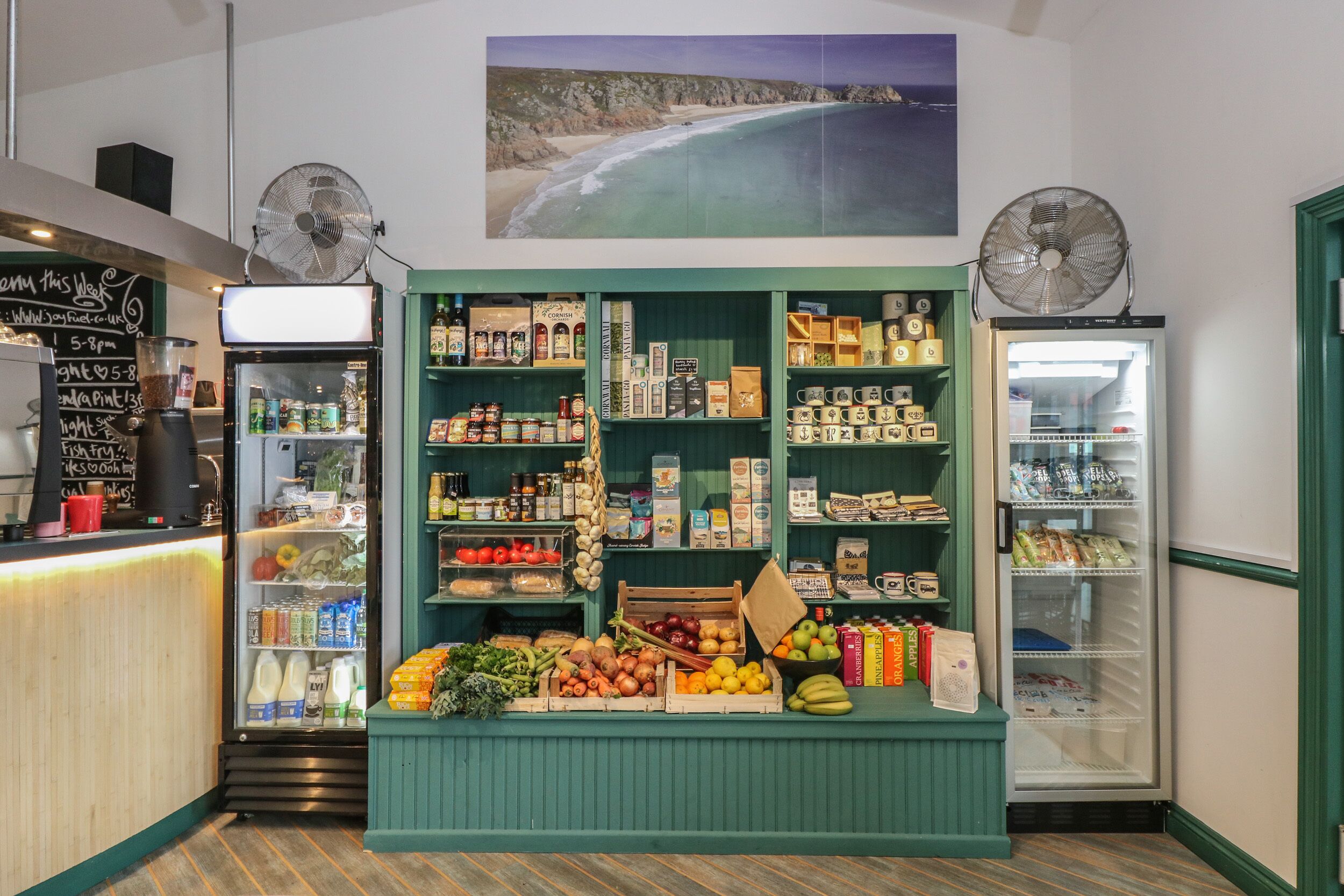A cafe interior with a coffee machine, a chalkboard menu, and a variety of food and drink items displayed on shelves and counters.