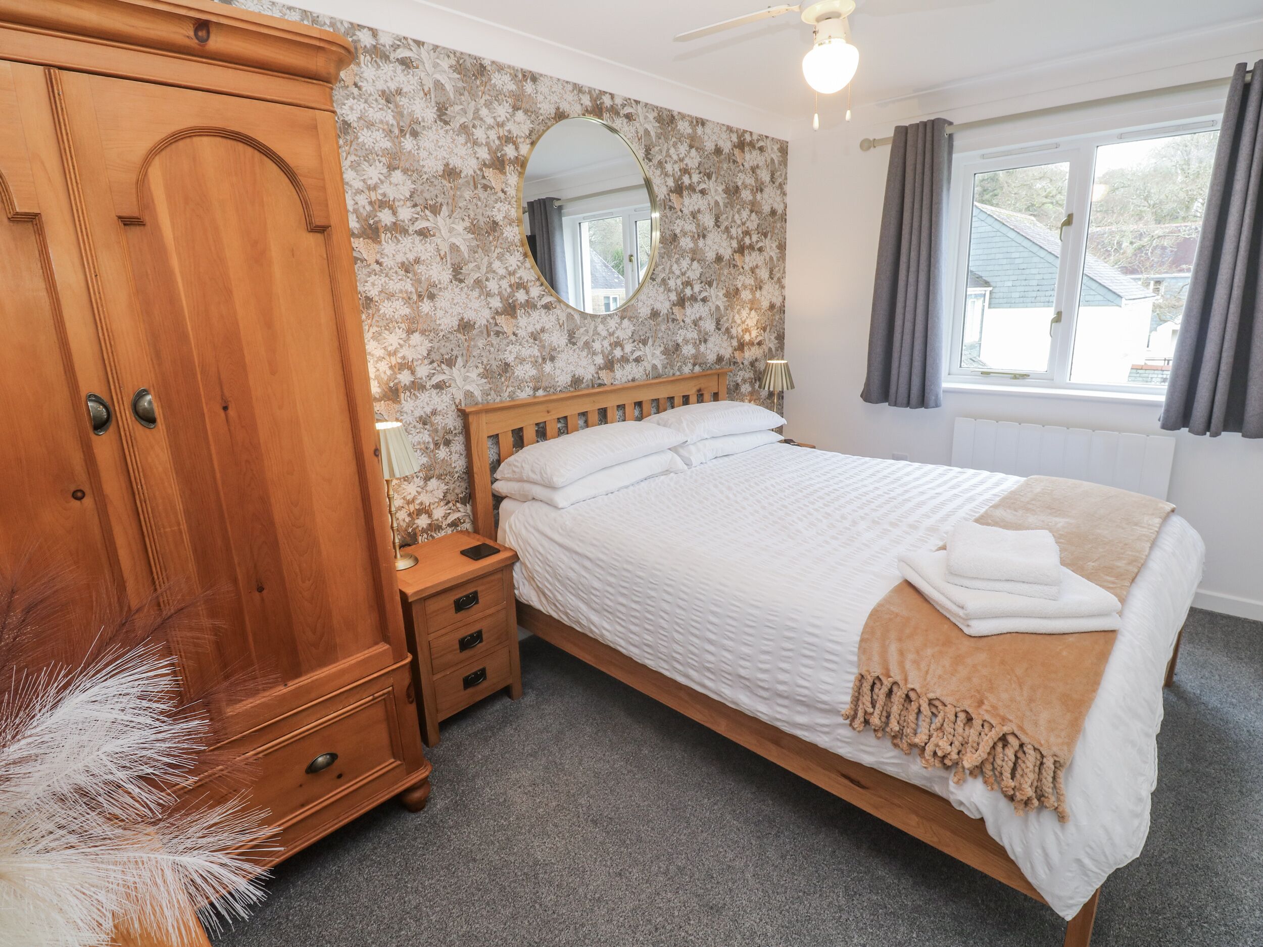A bedroom with a wooden wardrobe, a round mirror, a bed with white bedding, and a window with curtains.