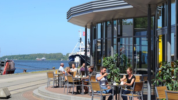 An outdoor cafe with tables and chairs, overlooking a body of water with boats and a ship in the background. - Hotel Am Kai 334 (Rostock)