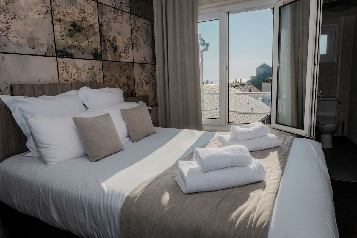 Ein Hotelzimmer mit einem großen Bett, Blick auf die Stadt durch eine Schiebetür und einem Badezimmer mit Toilette und Waschbecken.