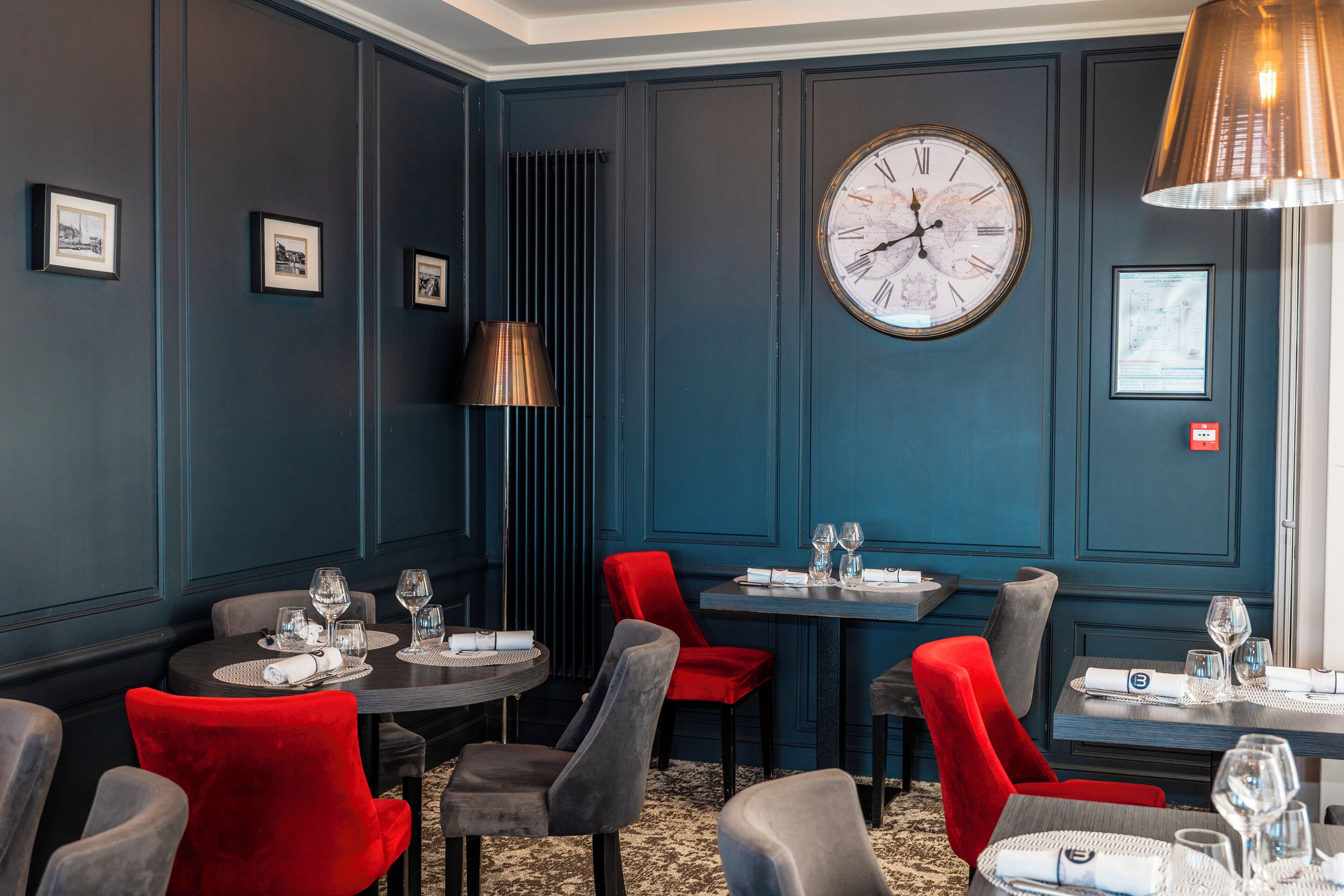 A dining area with dark blue walls, a large wall clock, and red and grey chairs.