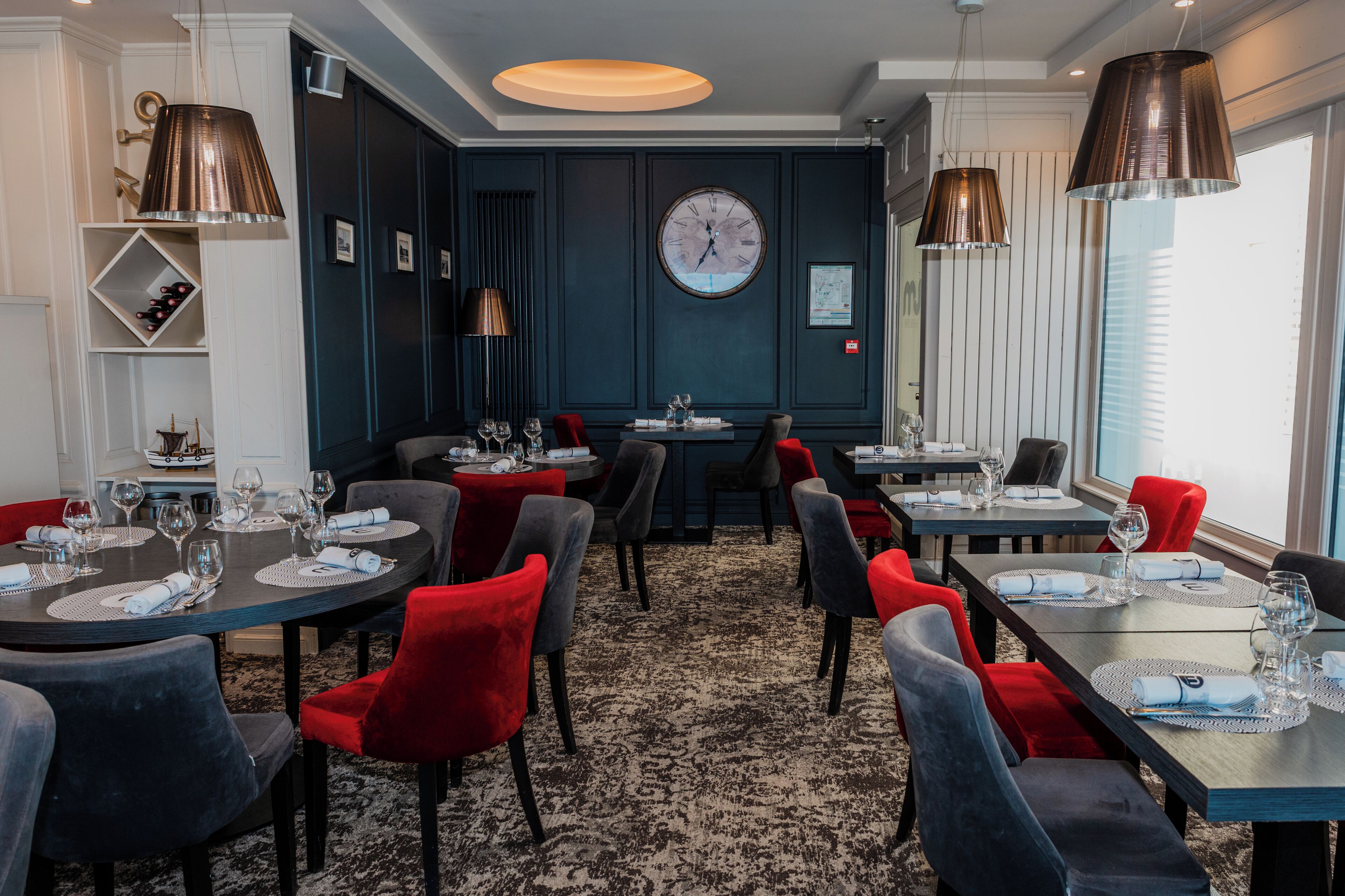 A dining area with multiple tables, red and grey chairs, and a large wall clock.