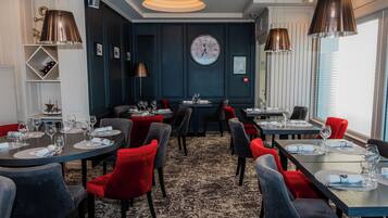 A dining area with multiple tables, red and grey chairs, and a large wall clock.