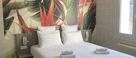 A neatly made bed with white linens and two folded towels, a nightstand with a lamp, and a wall with a tropical leaf pattern.