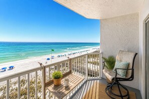 A balcony with a view of a beach, a chair, a table with a plant and a mug, and a railing. - Okaloosa Island Gulf Front Paradise Studio Condo (Fort Walton Beach)