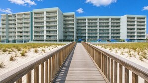 Un hotel frente a la playa con varios balcones, un paseo marítimo de madera que lleva a la entrada y un cielo azul despejado.