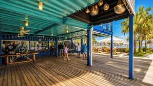Un espacio al aire libre techado con una terraza de madera, un bar de estilo azul y vistas a palmeras y un cuerpo de agua.