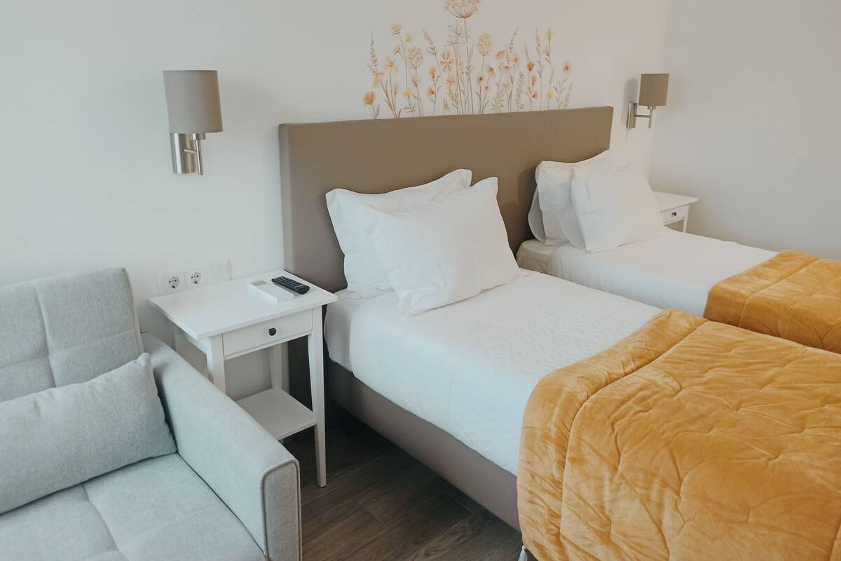 A hotel room with two beds, a sofa, a nightstand with a remote control, and wall-mounted lamps.