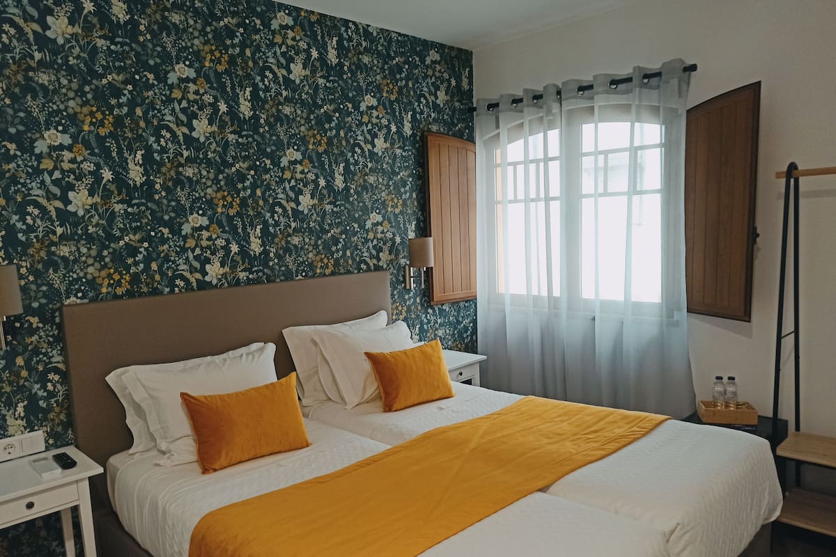A hotel room with a large bed, a nightstand, a lamp, a wooden wardrobe, and a window with sheer curtains.