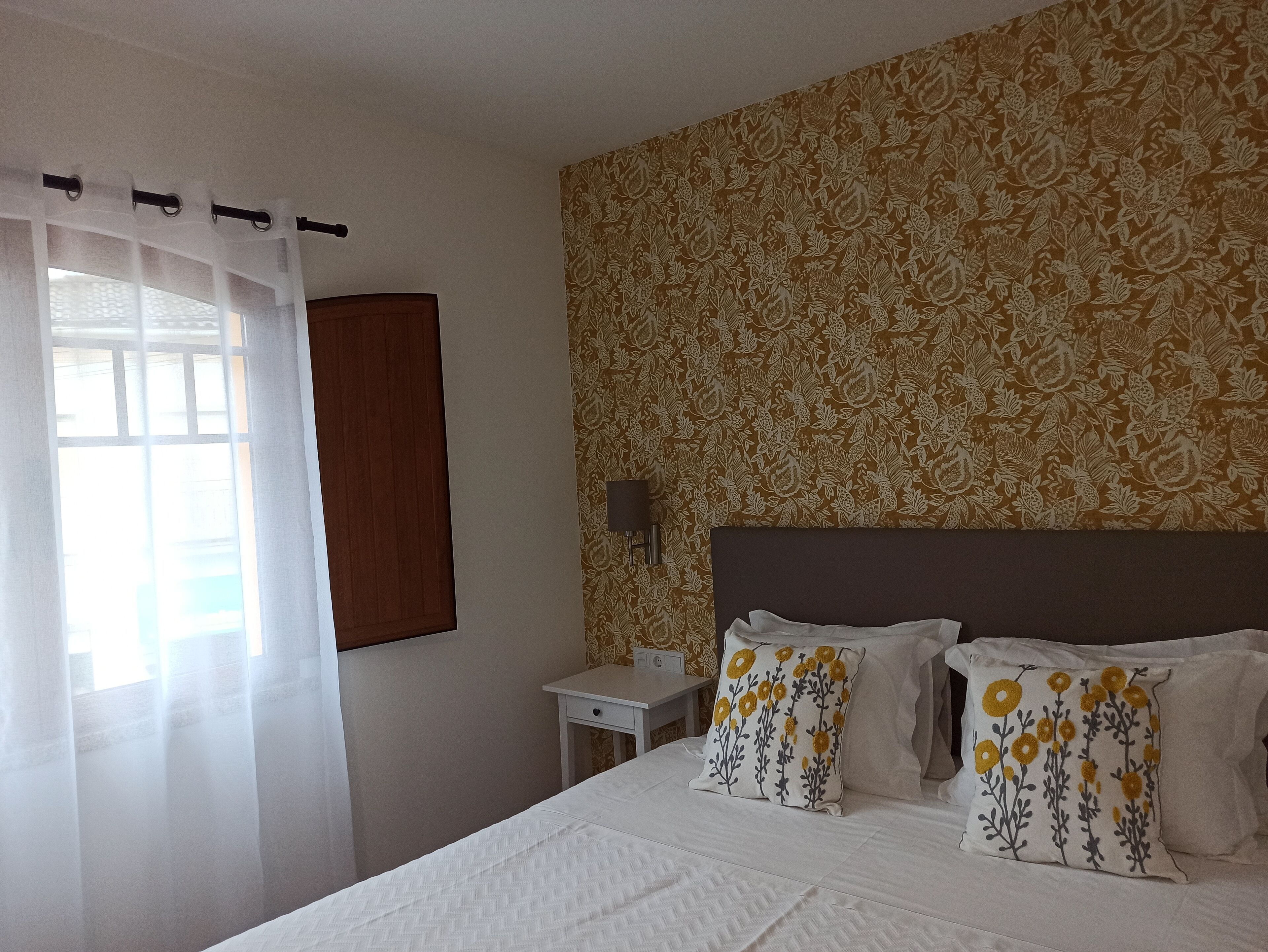 Een hotelkamer met een bed, witte gordijnen, een houten hoofdeinde, een nachtkastje en behang met een patroon.