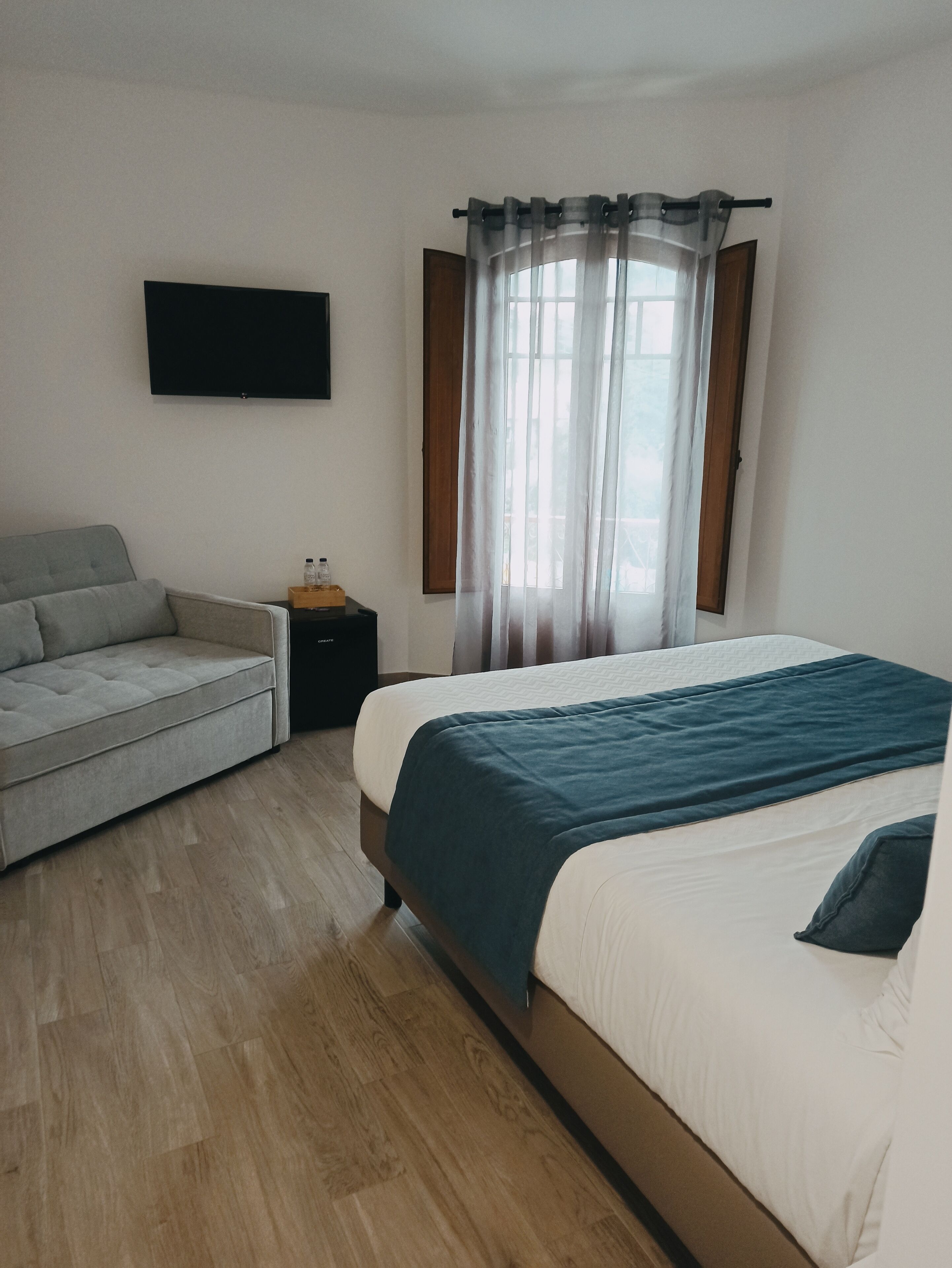 Quarto com cama, sofá, televisão na parede e janela com cortinas.