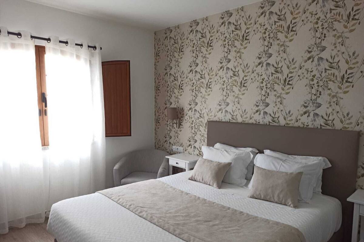 A bedroom with a large bed, a bedside table, a chair, and a window with curtains.