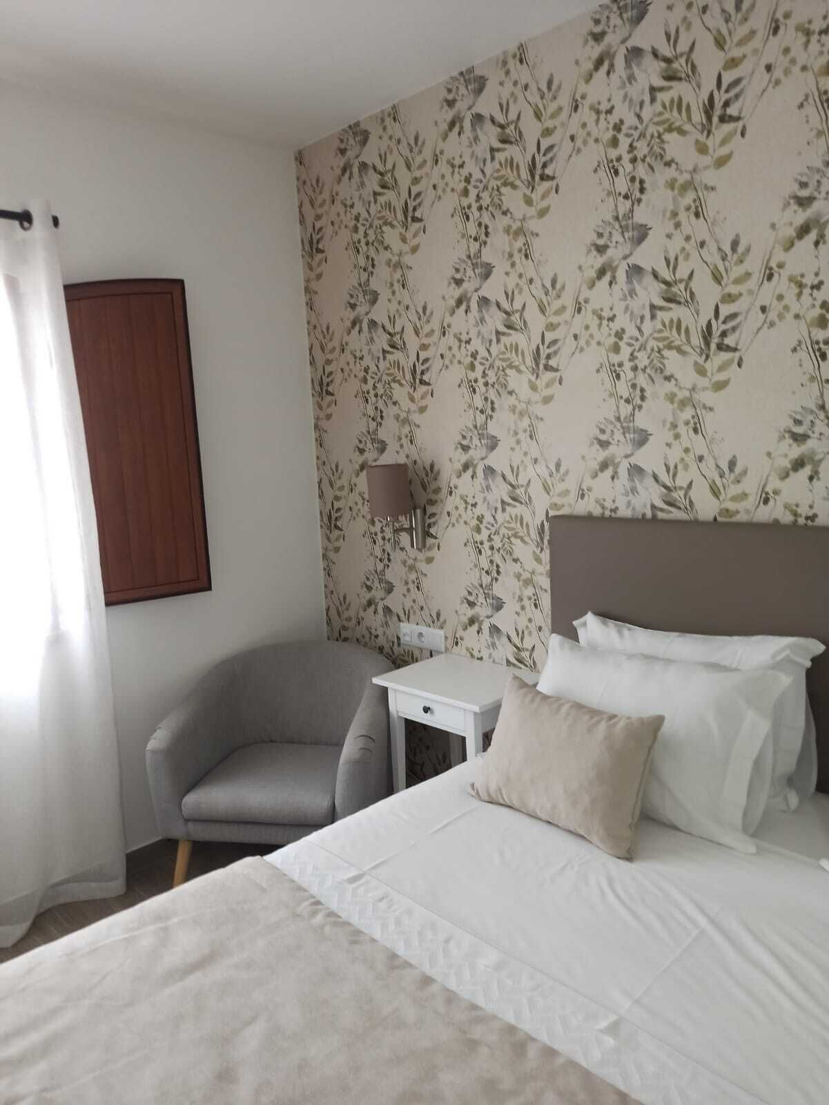 Una habitación de hotel con una cama, una silla, una mesita y un papel tapiz floral en la pared.