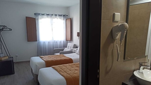 A hotel room with two beds, a bathroom with a sink and mirror, and a window with curtains. - Porto d'Abrigo - Alojamento Local (Sardoal)