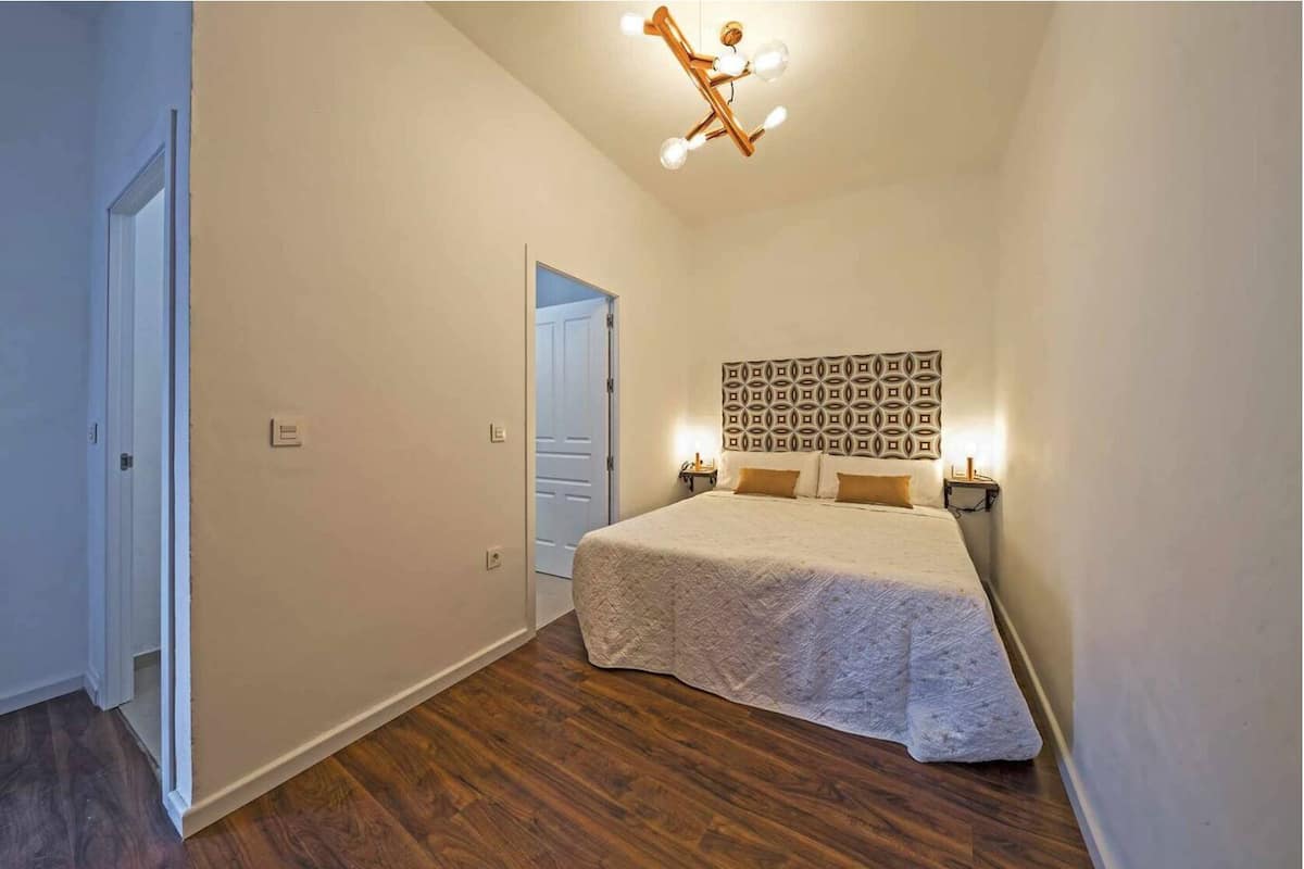 Un dormitorio con suelo de madera, una cama con cabecera estampada, lámparas de noche y una lámpara de techo.