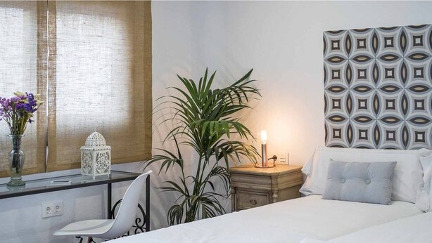 Un dormitorio con cama, mesita de noche, lámpara, planta en maceta y un panel decorativo en la pared.