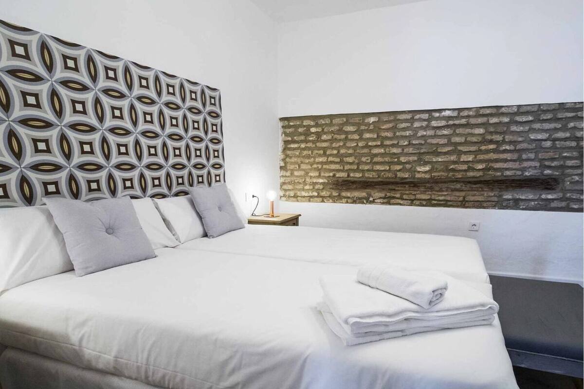 Una cama bien tendida con sábanas blancas y un papel tapiz estampado al fondo.