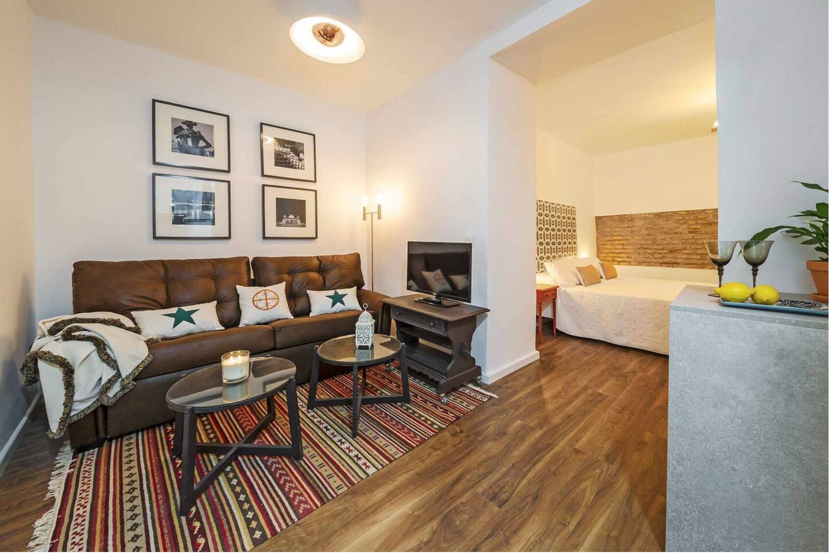 A modern living room with a brown leather sofa, a TV, and a small coffee table. There are framed pictures on the wall, a patterned rug, and a wooden floor.