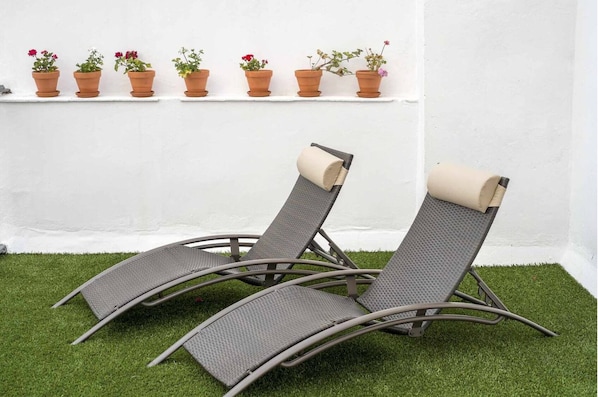 Two reclining chairs with a textured fabric and a beige cushion, placed on artificial grass against a white wall with a row of terracotta pots containing various flowers.
