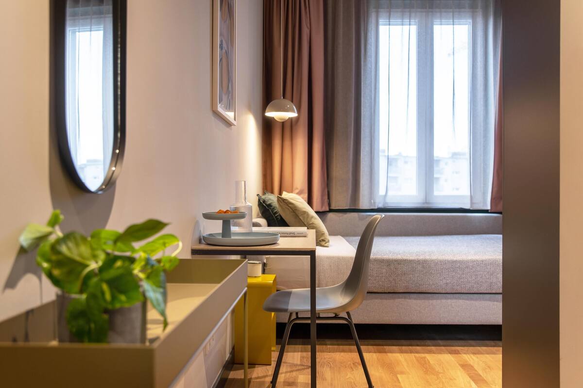 A modern room with a desk, a chair, a bed, a window with curtains, and a potted plant.