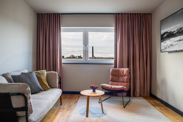 A modern living room with a sofa, a chair, a small table with a bowl of fruit, and a window with a cityscape view.