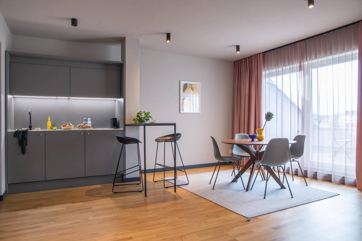 A modern kitchen with grey cabinets, a dining area with a table and chairs, and a large window with curtains.