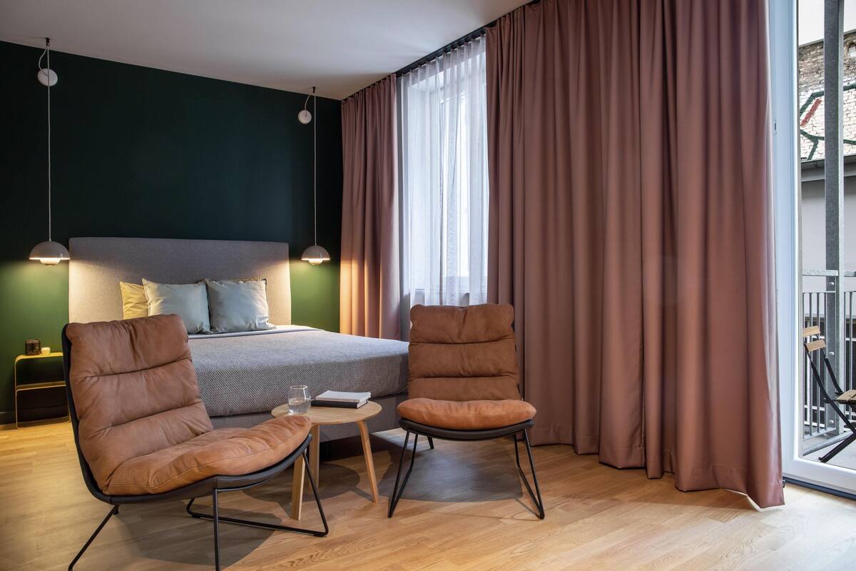 A modern hotel room with a bed, two chairs, a small table, and a large window with curtains.