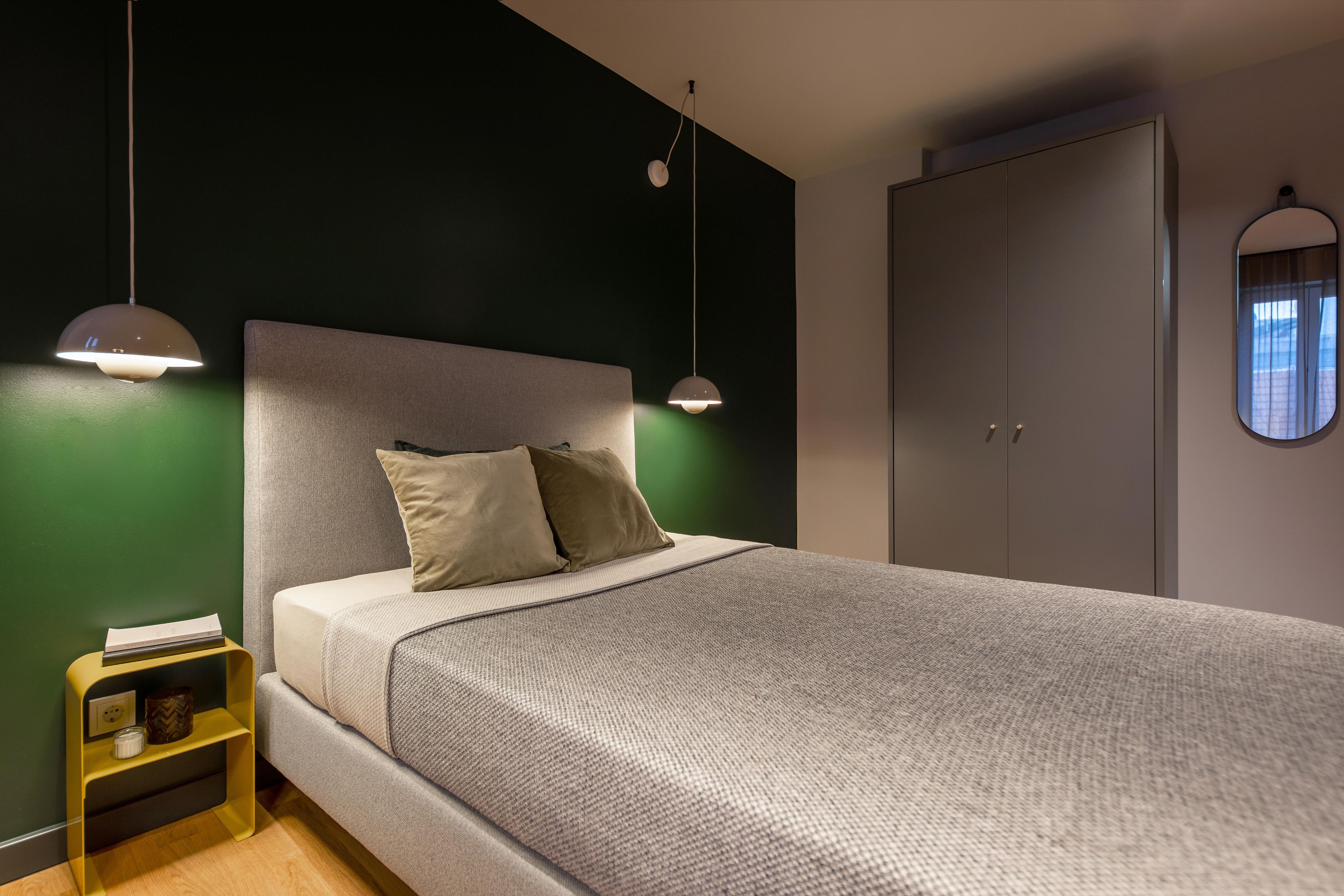 Un lit bien fait, avec une tête de lit grise et deux oreillers, une table de chevet avec une lampe et une armoire avec un miroir.