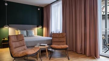 A modern hotel room with a bed, two armchairs, a small table, and a window with curtains.