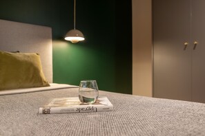 A glass of water on a magazine on a textured carpet, a hanging light, a green wall, and a bed with a yellow pillow.