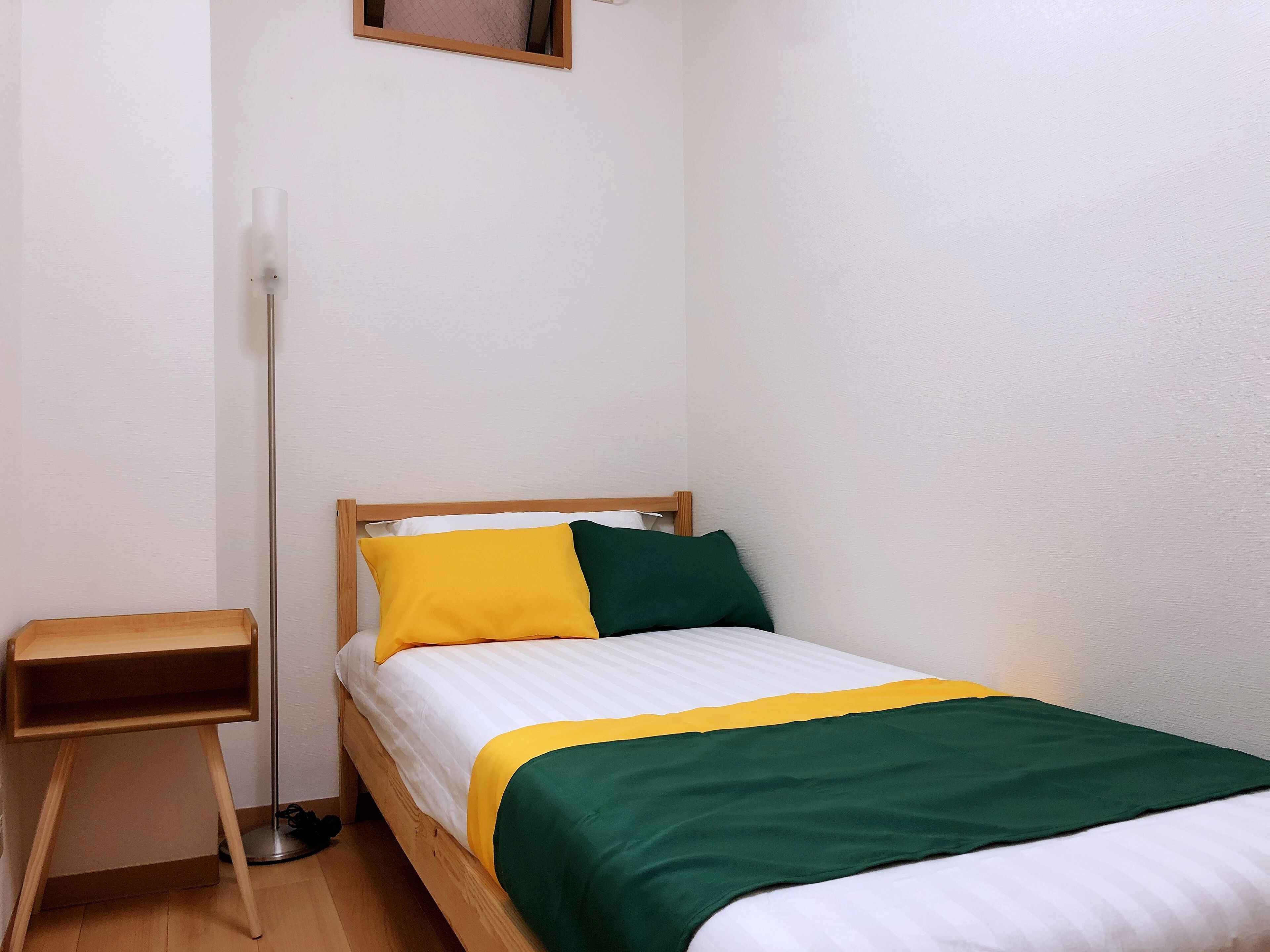 Un letto piccolo e ben rifatto con struttura in legno e materasso bianco, corredato da una coperta verde e gialla.