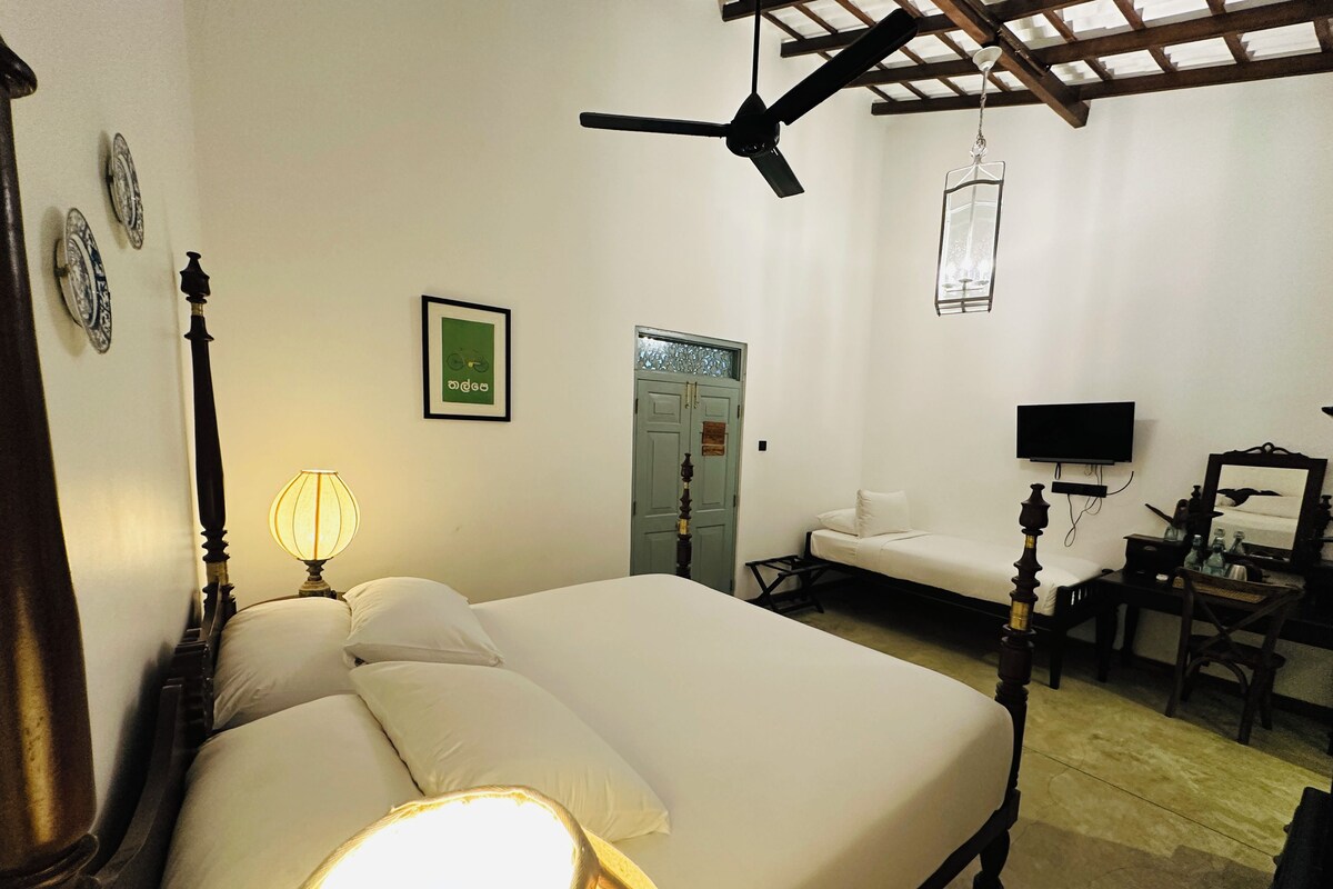 Quarto com cama, luminária de cabeceira, ventilador de teto, televisão e escrivaninha com cadeira.