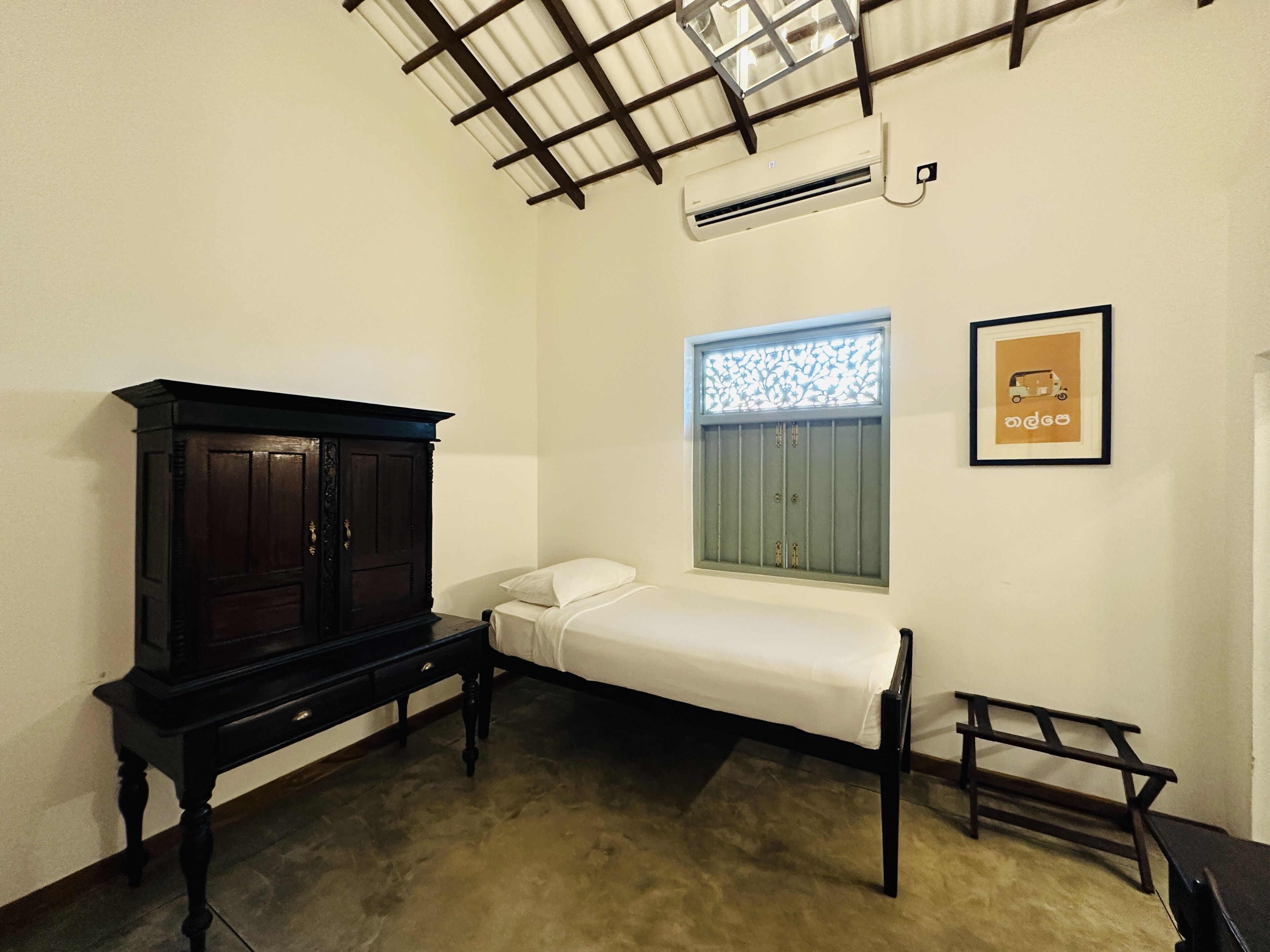 A small, single-bed room with a wooden wardrobe, a window with decorative bars, and a framed picture on the wall.