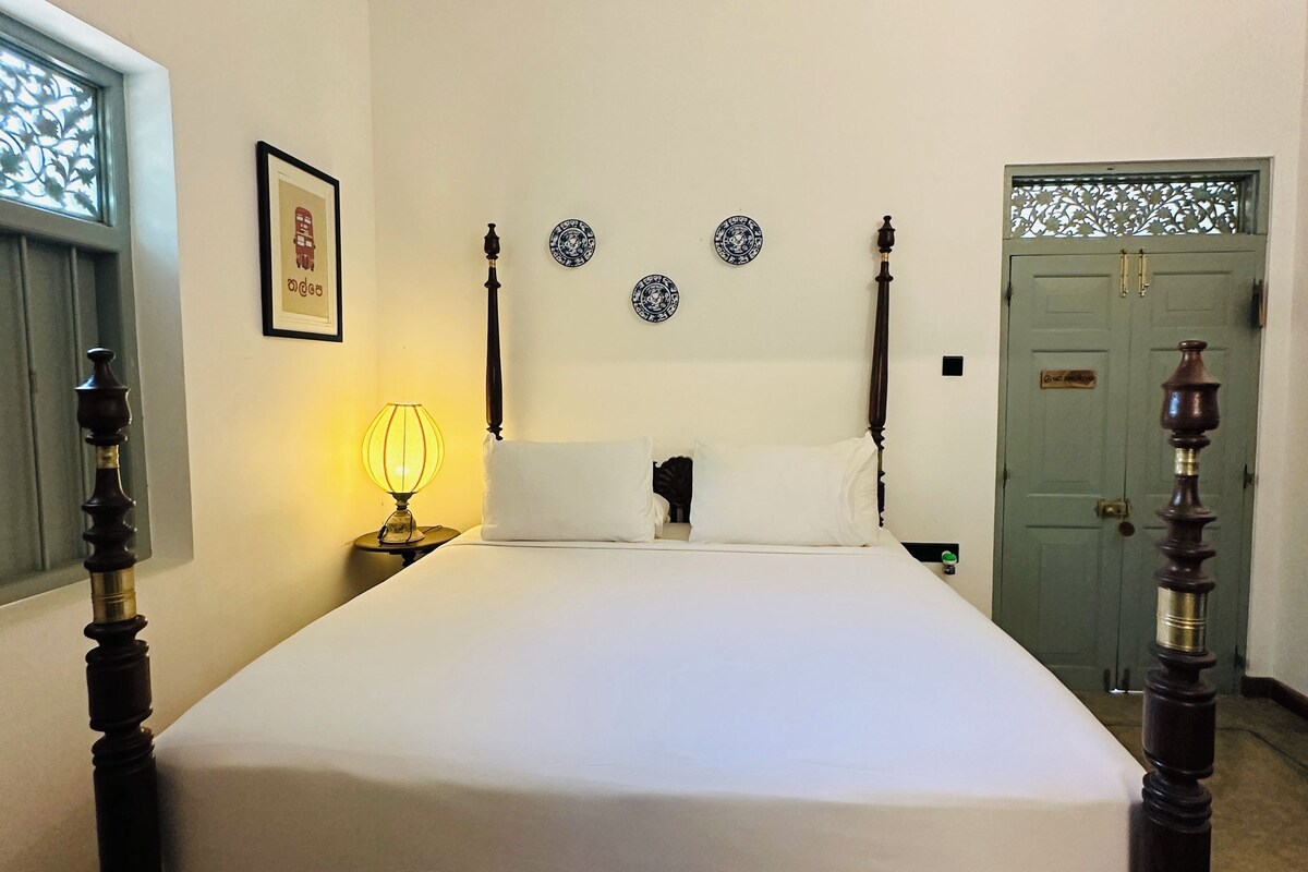 Una cama bien hecha en una habitación con cabecera de madera, un cuadro enmarcado en la pared y una puerta verde con un ventanal decorativo.
