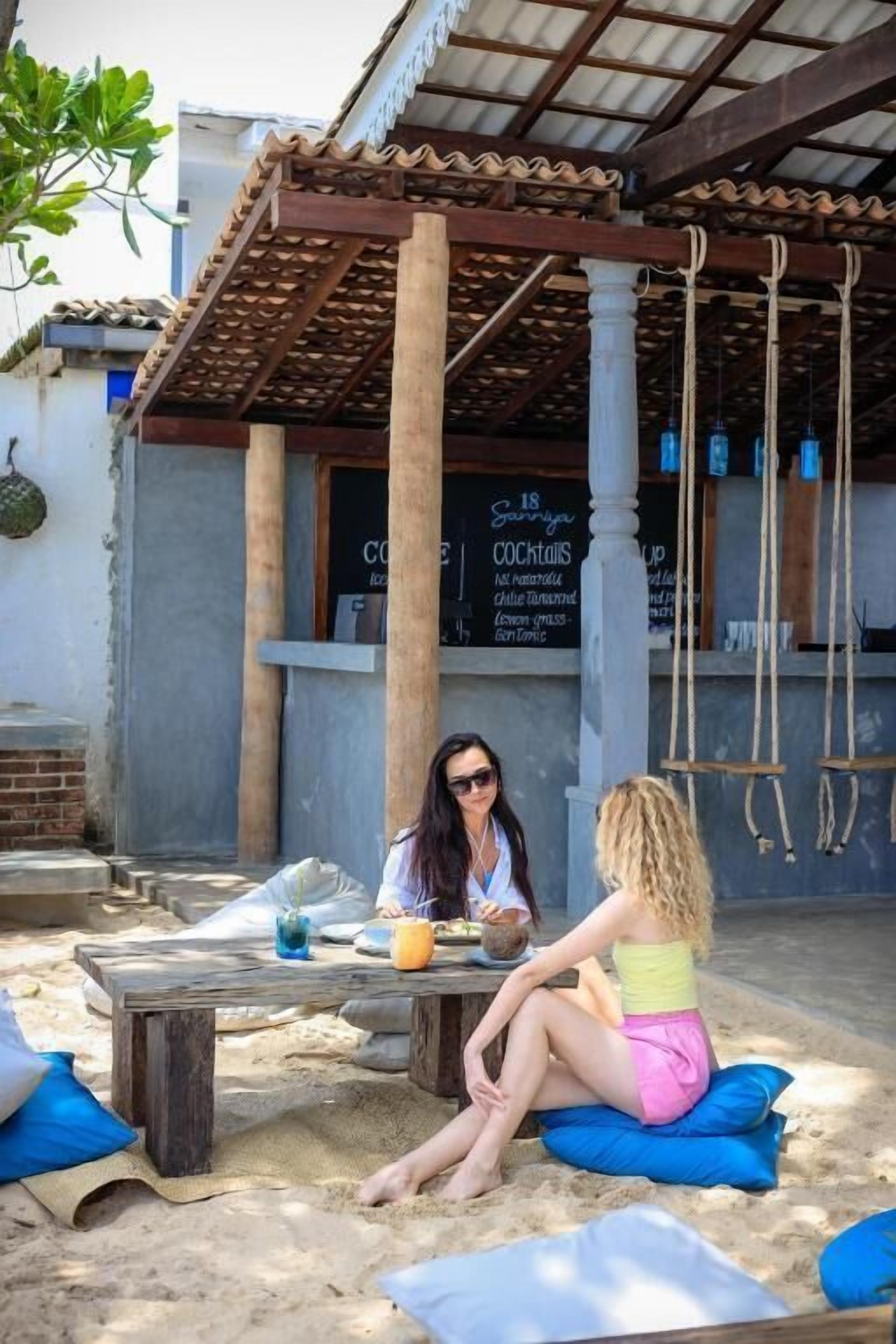 Two people sitting at a wooden table with a drink and a menu in a covered outdoor area with columns and a hanging light fixture.