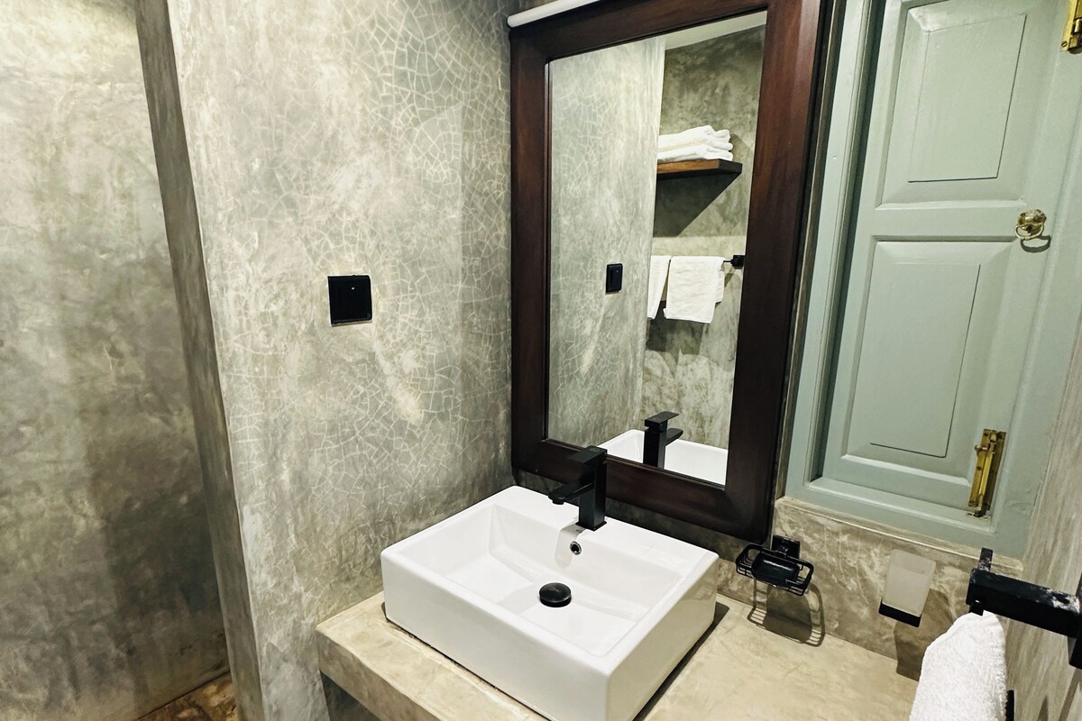 Un baño con un lavamanos blanco, un espejo, un perchero para toallas y una puerta.