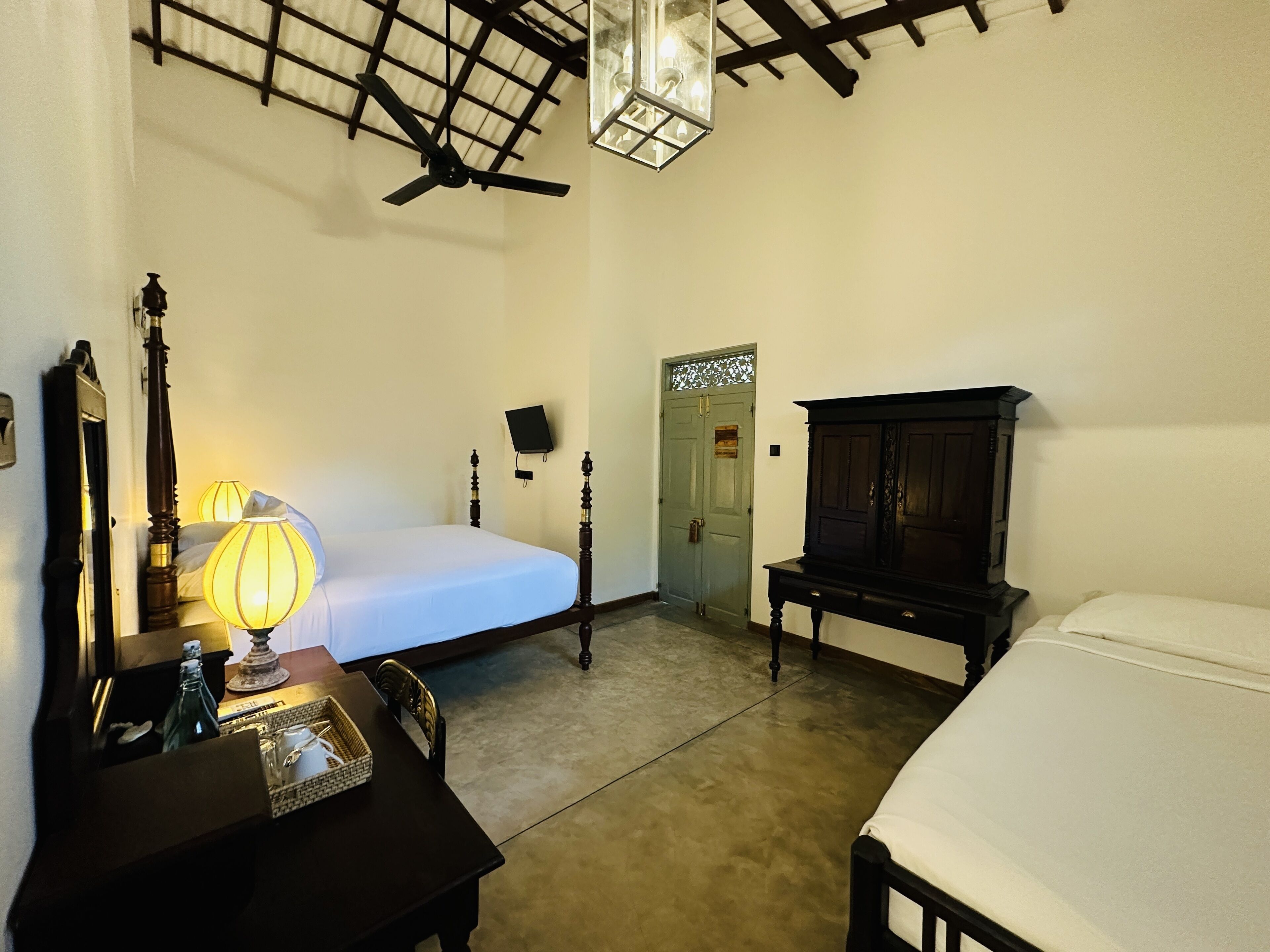 A room with two beds, a desk with a lamp, a wooden cabinet, and a ceiling fan.