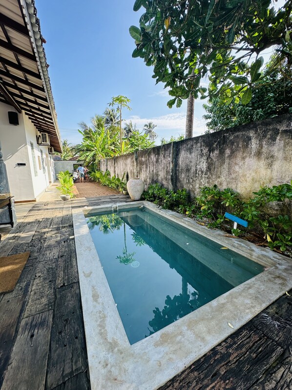 Une piscine dotée d’une terrasse en bois, entourée de verdure et bordée d’un mur en béton.