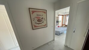 Un couloir carrelé mène à une chambre à coucher où se trouvent un lit et un fauteuil rouge.