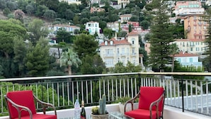 Un balcon avec deux chaises rouges, une table en verre et une bouteille d’eau Evian.