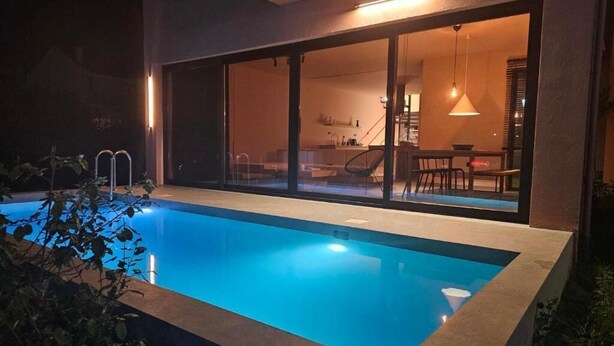 Una piscina con recinto in vetro, una zona pranzo con tavolo e sedie, e una scala che conduce a un piano superiore.