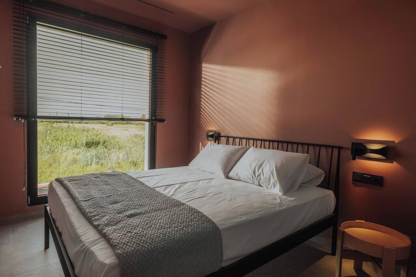 A bedroom with a bed, a window with blinds, a bedside table, and a wall-mounted light fixture.