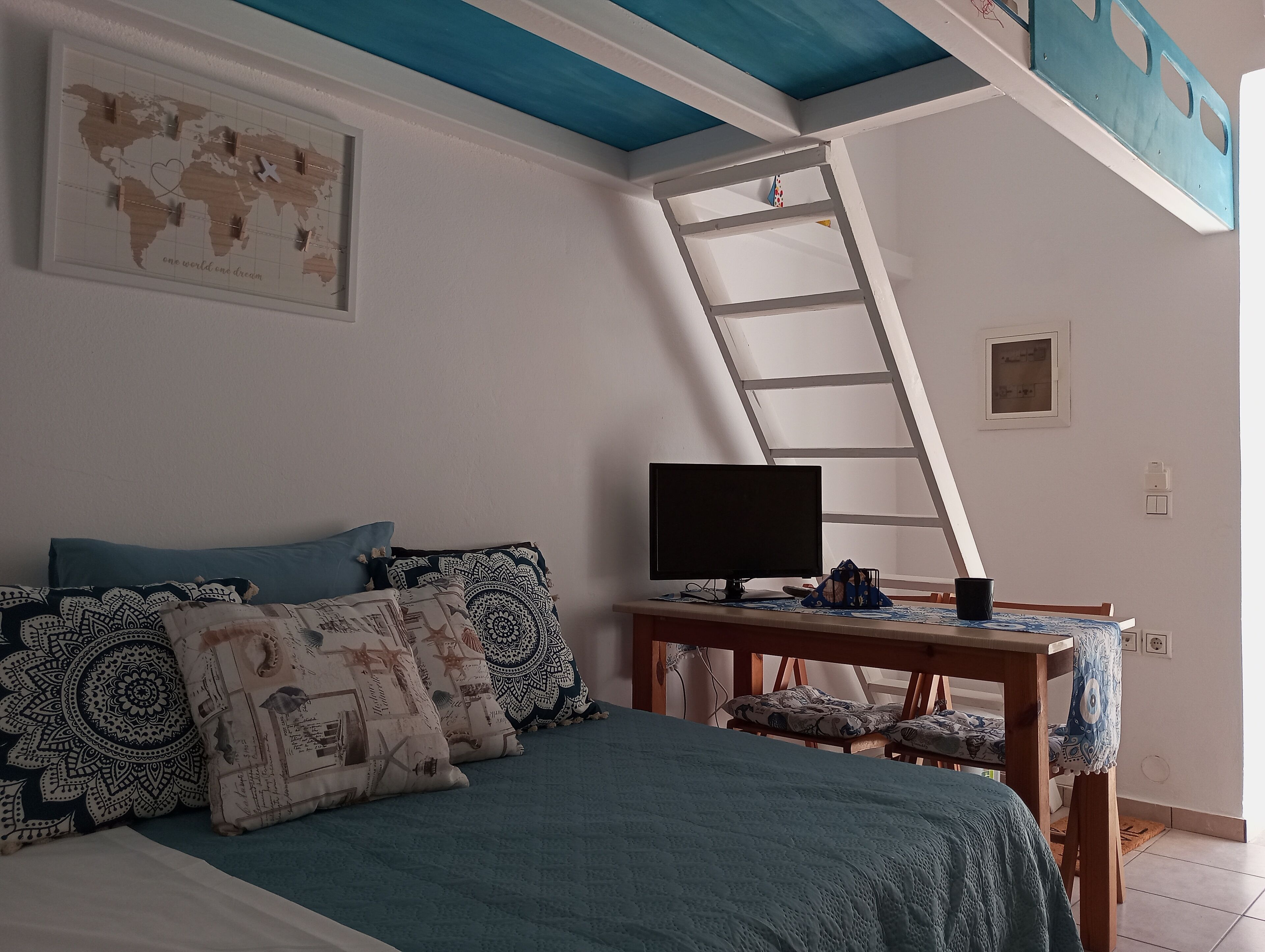 Ein Schlafzimmer mit einem Bett, Kissen, einem Schreibtisch mit Computer, einer Weltkarte an der Wand und einem Oberlicht.