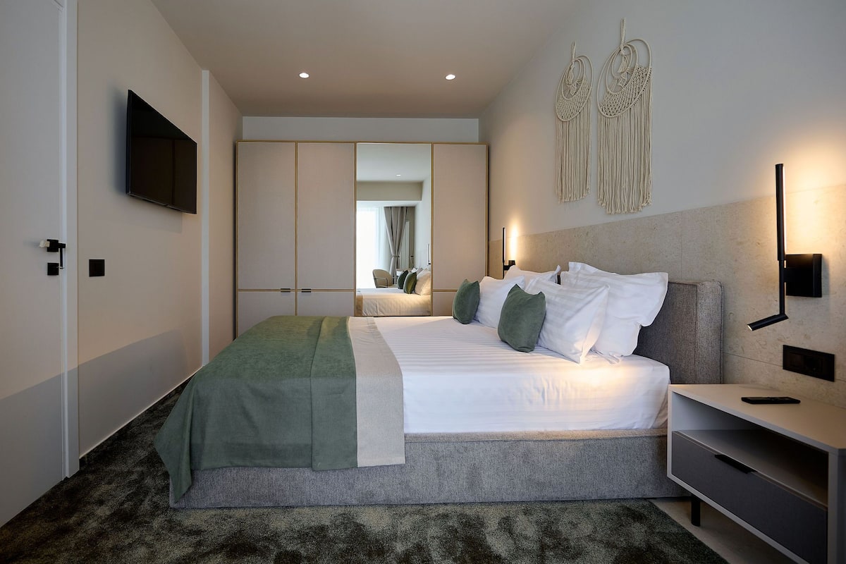 Una habitación de hotel moderna con una cama grande, una mesita de noche, un televisor montado en la pared y un armario con puertas correderas.