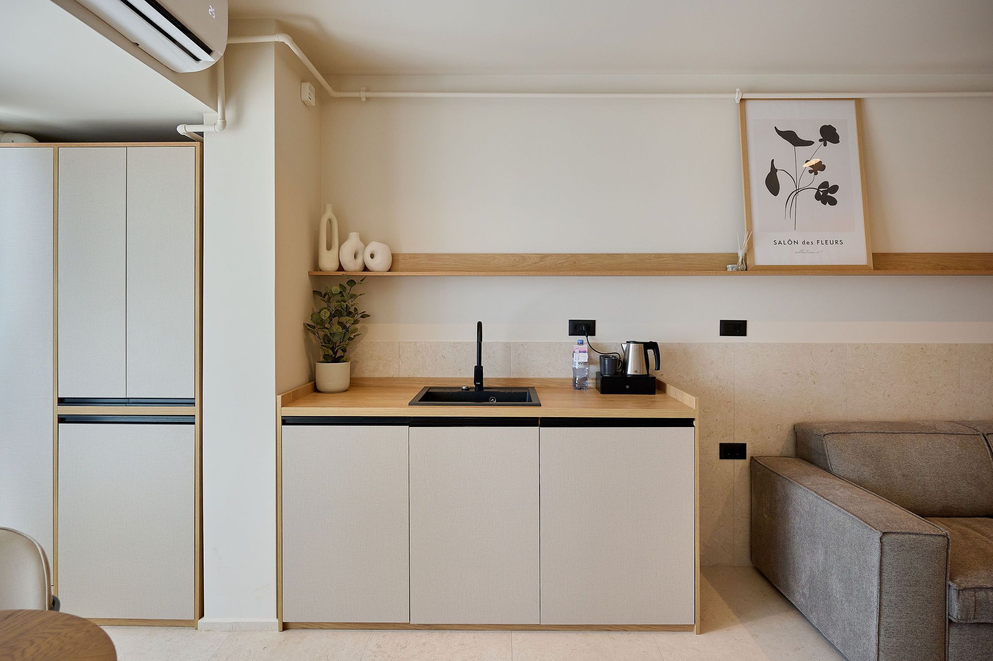 Una cocina moderna con lavavajillas integrado, un lavamanos y una cafetera. Hay una repisa de madera sobre la encimera con objetos decorativos y un cuadro enmarcado.