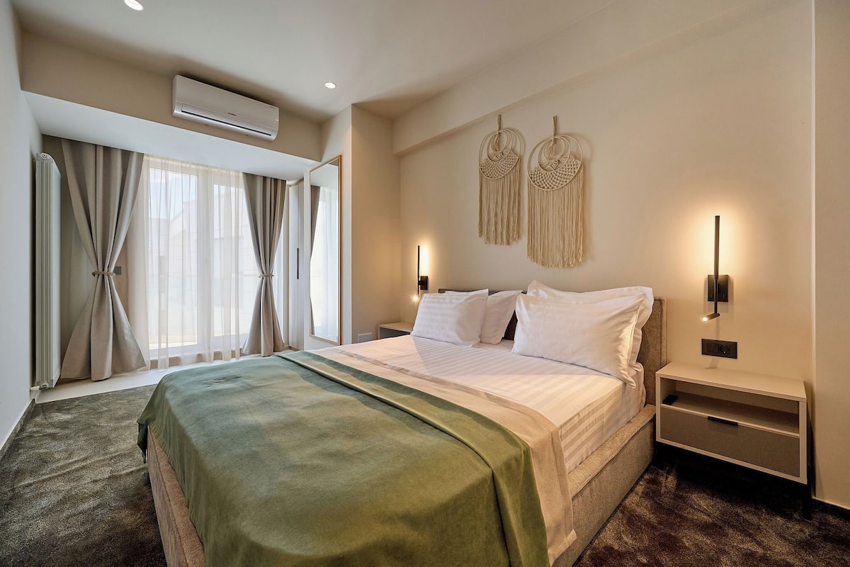 Una habitación de hotel con cama, mesitas de noche, aire acondicionado y un ventanal con cortinas.