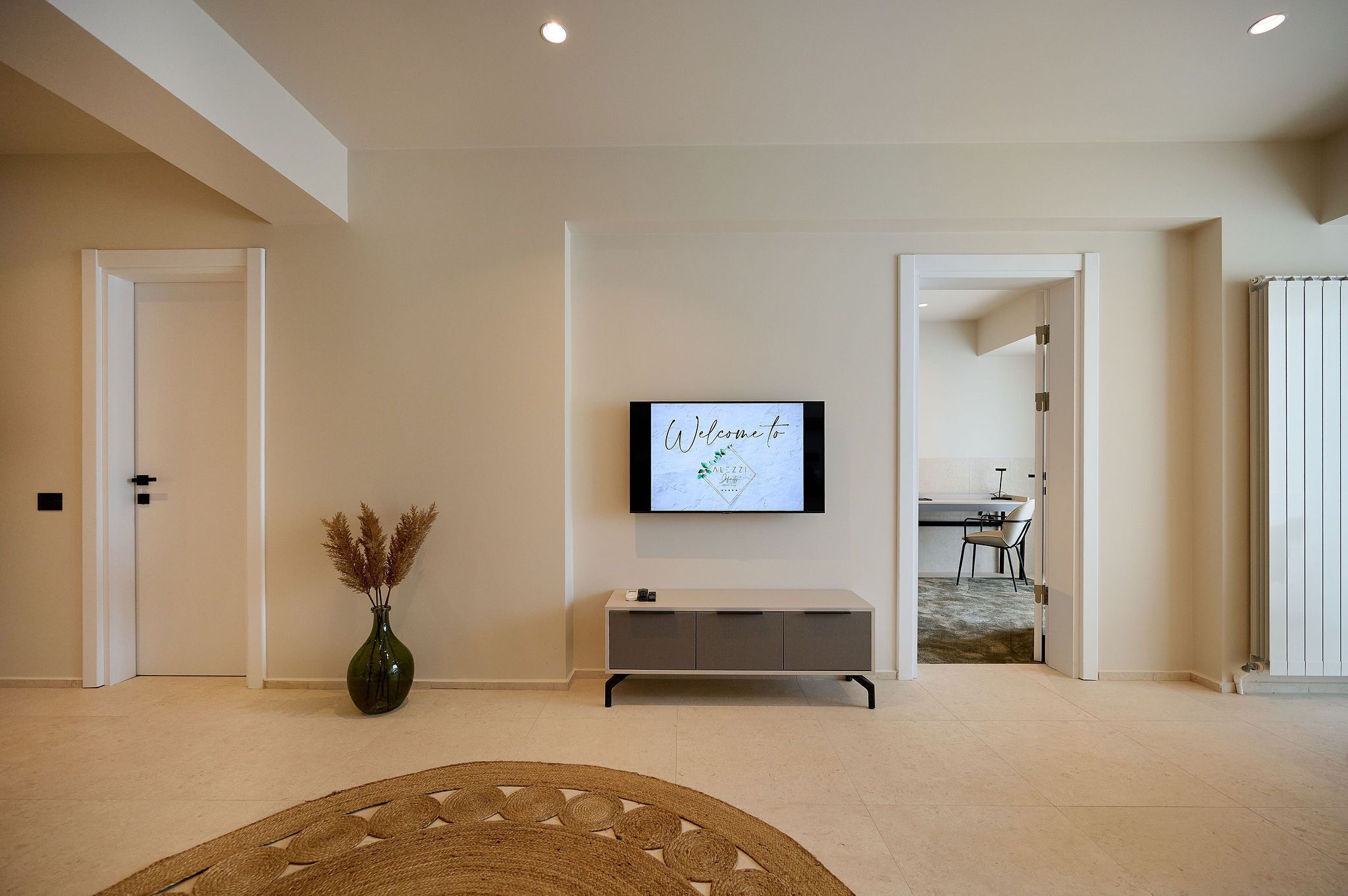 Un salon moderne avec un téléviseur à écran plat fixé au mur, une console grise et un vase décoratif contenant des plantes séchées.