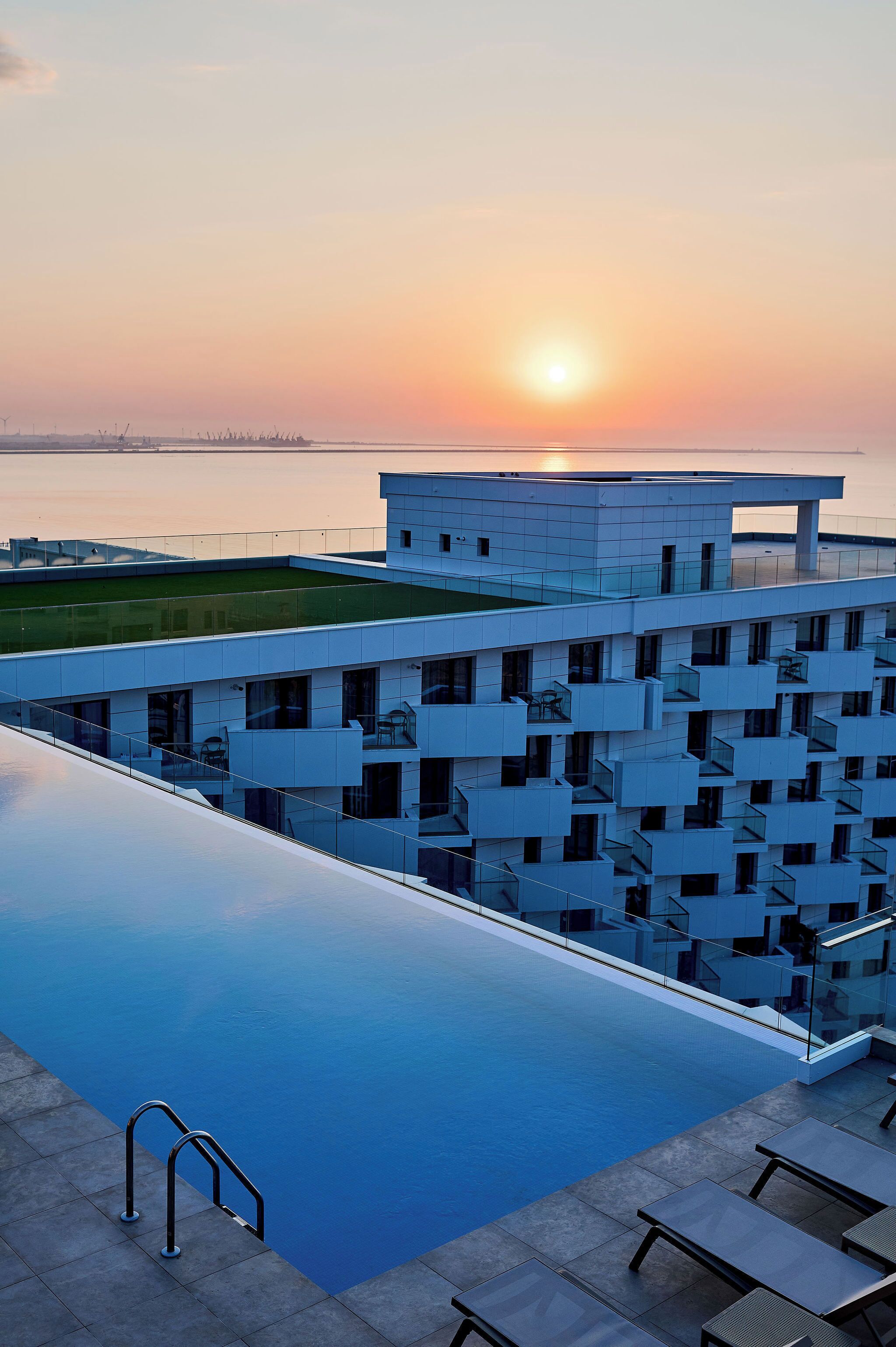 Ein modernes Hotel mit einem Dachpool im Infinity-Stil, einer Glasbrüstung und Blick auf den Sonnenuntergang über dem Wasser.